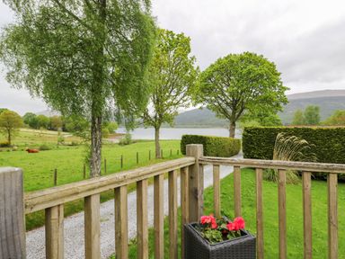 View of a wooden fence overlooking a gravel path green grass trees and a lake at The Four Oaks Chalet in Blaich near Fort William