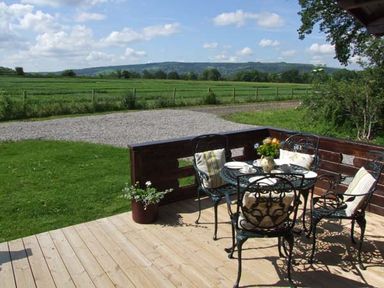 An outdoor wooden deck with a metal table and four chairs with cushions a potted plant and a countryside field view at Cropvale Lodge in Cropthorne near Pershore