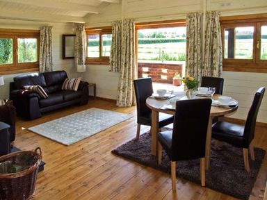 A dining table with four chairs and a vase of flowers near a black sofa with cushions and windows at Cropvale Lodge in Cropthorne near Pershore
