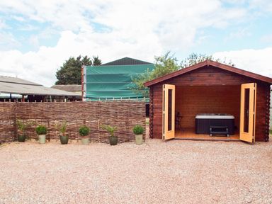 A wooden shed with open doors revealing a hot tub inside and potted plants lined up along a woven fence at Cropvale Lodge in Cropthorne near Pershore