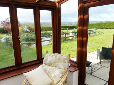 A conservatory with a cushioned chair and pillow overlooking a garden and patio at The Old Granary in Polyphant near Launceston