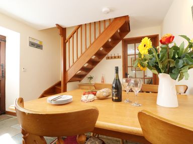 A dining room with wooden table and chairs flowers wine bread and cheese on the table and wooden stairs in the background at The Old Granary in Polyphant near Launceston
