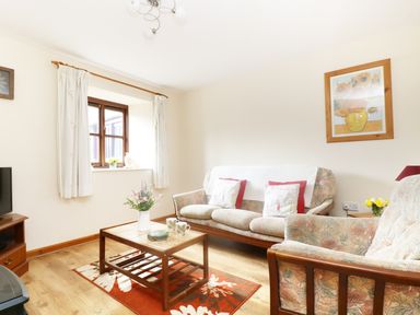 A living room with a floral sofa set a wooden coffee table with flowers and mugs a window with white curtains and a painting on the wall at The Old Granary in Polyphant near Launceston