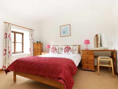 A bedroom with a double bed red quilt floral curtains wooden furniture and a dressing table at The Old Granary in Polyphant near Launceston