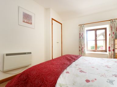 A bedroom with a bed covered in floral bedding a red quilt a window with floral curtains a wooden dresser a wall heater and a built in closet at The Old Granary in Polyphant near Launceston
