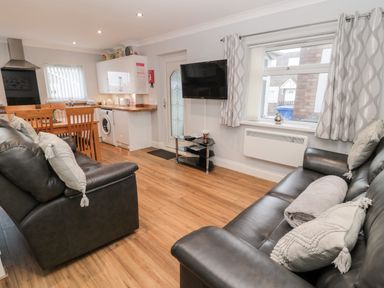 A living area with black leather sofas a wall-mounted TV wooden dining table and chairs and a kitchen with white cabinets at Cheviot View in Beadnell