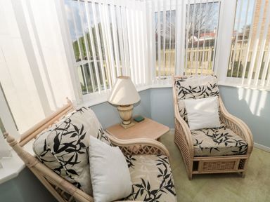 A sunroom with two wicker chairs with cushions a small table with a lamp and vertical blinds at Cheviot View in Beadnell