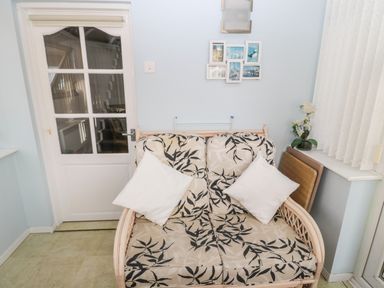 A seating area with a patterned cushioned loveseat white pillows a glass paneled door a set of pictures on the wall and a window with blinds and a flower pot on the sill at Cheviot View in Beadnell