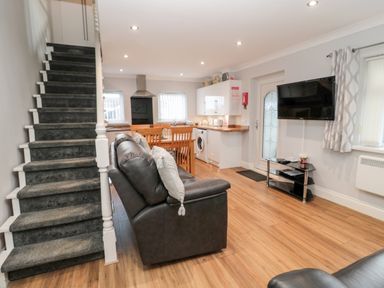An open plan living room and kitchen with a black leather sofa a wooden dining table stairs and a wall mounted TV at Cheviot View in Beadnell
