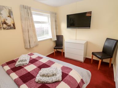 A bedroom with a bed covered by a red and white check blanket towels on the bed two black chairs a white dresser and a wall mounted flat screen TV at Cheviot View in Beadnell