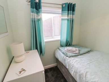 A small bedroom with a single bed covered with a light blue bedspread and a white bedside table with a lamp near a window with blue striped curtains at Cheviot View in Beadnell