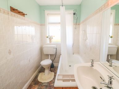 A bathroom with a toilet a bathtub with shower curtain a sink and a window with flowers on the sill at Kates Cottage in Knightstown Valentia Island County Kerry