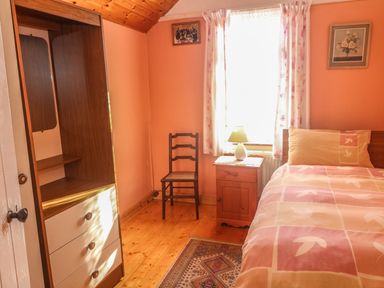 A bedroom with a bed bedside table lamp chair wardrobe and window with curtains at Kates Cottage in Knightstown Valentia Island County Kerry