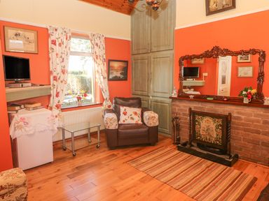 A living room with a brown armchair, floral curtains, a TV, a glass table, a fireplace with a large wooden framed mirror above it at Kates Cottage Knightstown Valentia Island County Kerry