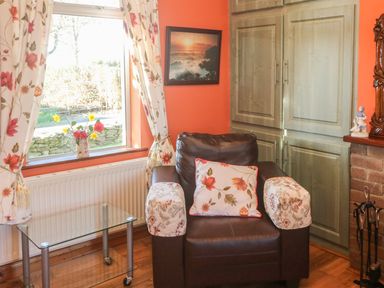 A room corner with a brown armchair with floral covers and a cushion a glass table and a window with floral curtains at Kate's Cottage Knightstown Valentia Island County Kerry