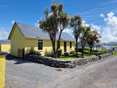 A yellow house with a stone wall and palm trees along a gravel road at Kates Cottage in Knightstown Valentia Island County Kerry