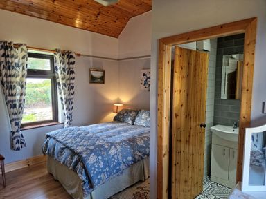 A bedroom with a bed with blue floral bedding a window with blue and white curtains and a doorway to a bathroom at Kates Cottage in Knightstown Valentia Island County Kerry