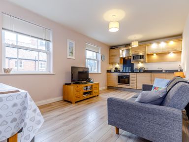 A living area with a gray sofa a wooden TV stand with a television two windows and a kitchen with wooden cabinets at Sunrise The Bay near Filey
