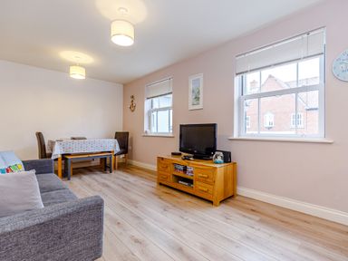 A living room with a gray sofa a wooden TV stand with a television a dining table with chairs and two windows at Sunrise The Bay near Filey