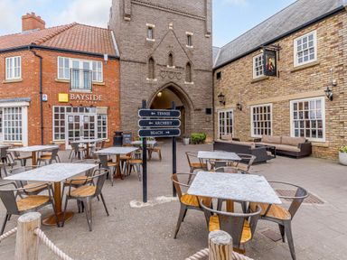 Outdoor seating area with tables and chairs in front of brick buildings including Bayside Kitchen at Sunrise The Bay near Filey