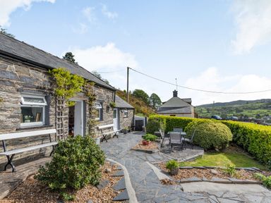 An outdoor patio with benches and a table with chairs beside a stone building at Henrhiw Bach in Penmachno