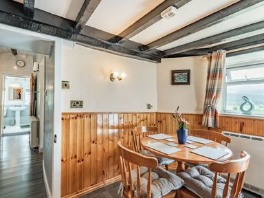 A dining area with a round wooden table and four chairs with cushions and a window with checkered curtains at Henrhiw Bach in Penmachno