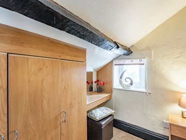 A corner of a room with a wooden wardrobe a small desk with a mirror vases with red flowers a window with a decorative ornament a lamp on a side table and a stool with a cushion at Henrhiw Bach in Penmachno