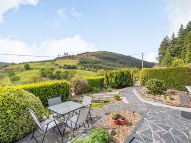 An outdoor patio with a glass table and four chairs surrounded by bushes and a stone walkway with hills and trees in the background at Henrhiw Bach in Penmachno