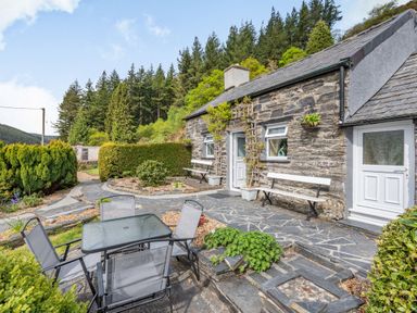 An outdoor patio area with a glass table and four chairs in front of a stone cottage with two white doors and two benches at Henrhiw Bach in Penmachno