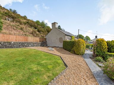 A yard with gravel and grass next to a stone house with a hill in the background at Henrhiw Bach in Penmachno