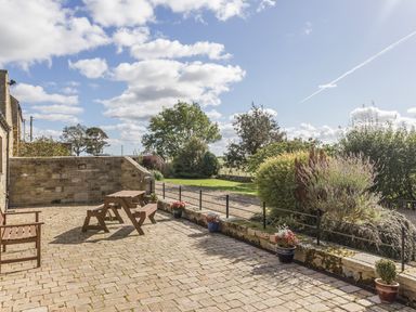 A stone patio with wooden bench and table set overlooking garden and trees at Ridge Cottage in Whitridge