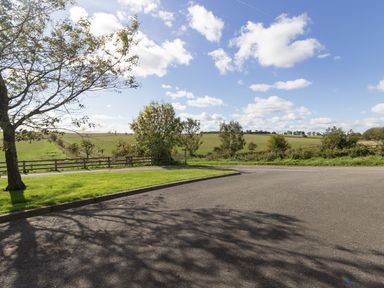 A paved area with grass and trees next to open fields under a partly cloudy sky at Ridge Cottage in Whitridge