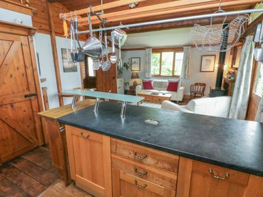 A kitchen with hanging cooking utensils and a black countertop overlooking a living room with white sofas and a wood stove at Timbertwig Lodge in Lamphey near Pembroke