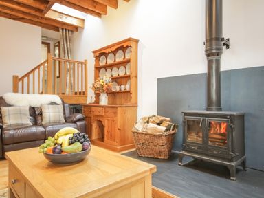 A living room with a wood-burning stove a basket of firewood a leather sofa a wooden cabinet with plates and mugs and a wooden coffee table with a fruit bowl at Pendre Uchaf in Melin-y-Wig near Ruthin