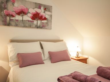 A bedroom with a bed white pillows pink cushions folded towels bedside table with lamp and flower painting at Pendre Uchaf in Melin-y-Wig near Ruthin