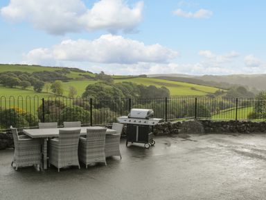 An outdoor patio with a dining table and chairs and a barbecue grill overlooking green hills and trees at Pendre Uchaf in Melin-y-Wig near Ruthin