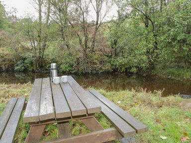 A wooden picnic table with two cups and a thermos by a river surrounded by trees at Pendre Uchaf in Melin-y-Wig near Ruthin