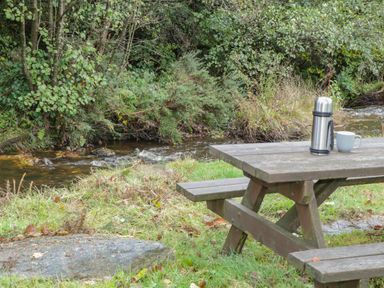 A wooden picnic table with a thermos and cup beside a small stream with trees and grass at Pendre Uchaf in Melin-y-Wig near Ruthin