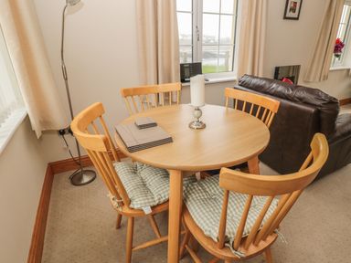 A dining area with a round wooden table and four chairs with cushions near a window and a brown leather sofa at White Rock Cottage in Beadnell