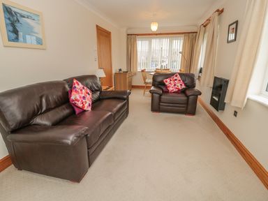 A living room with a leather sofa and armchair with pink cushions and a table by windows at White Rock Cottage in Beadnell