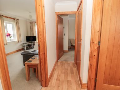 A hallway with wooden floor and doors leading to a living room with a sofa and television and a room with a chair at White Rock Cottage in Beadnell