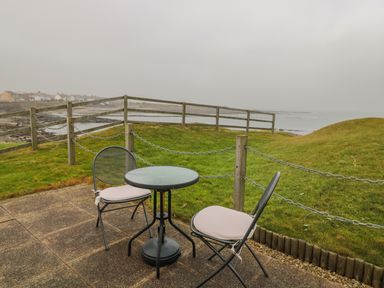 A patio with two metal chairs and a round glass table overlooking a grassy area and coastal view at White Rock Cottage in Beadnell