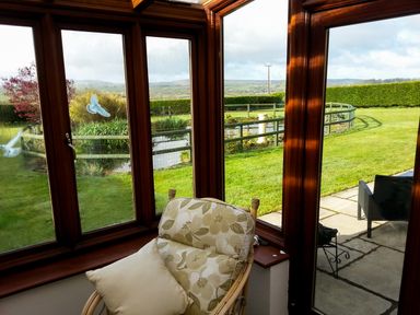 A conservatory with a cushioned chair and pillow overlooking a garden with a fenced pond and lawn at The Shippon Near Launceston