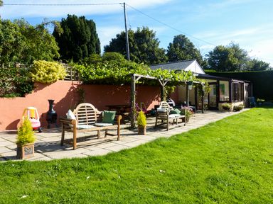An outdoor patio with wooden benches and a pergola covered with plants beside a green lawn at The Shippon Near Launceston