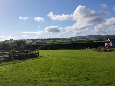 A large green lawn with a wooden fence and a small blue playhouse with a slide near a hedge and rolling hills in the background at The Shippon Near Launceston