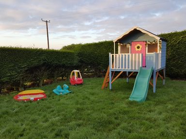 A backyard with a children's playhouse with slide a small sandbox a blue rocking toy and a red toy car