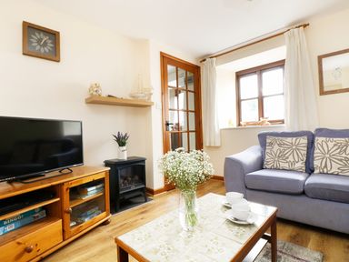 A living room with a blue sofa a wooden cabinet a TV a glass door a table with flowers and cups at The Shippon Near Launceston