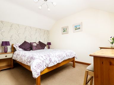 A bedroom with a wooden bed with purple patterned bedding and pillows bedside tables with purple lamps a wooden desk with a chair and two framed pictures on the wall at The Shippon Near Launceston