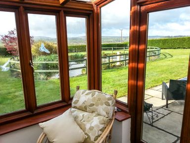 A seating area with a cushioned chair inside a sunroom with wooden framed windows looking out to a grassy garden with a pond and fencing at The Wagon House in Polyphant near Launceston
