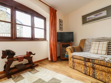 A corner room with a patterned sofa with a pillow and blanket a wooden rocking horse a TV on a wooden cabinet and large window with orange curtain at The Wagon House in Polyphant near Launceston